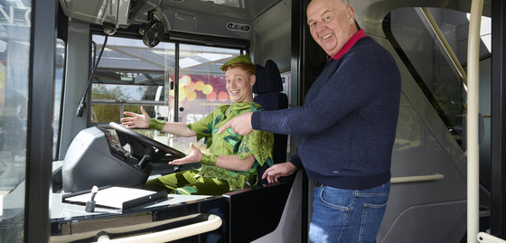 Peter Pan is sat at the driver's seat on a D&E Coach, with Donald from the company stood beside him, pointing to the wheel.